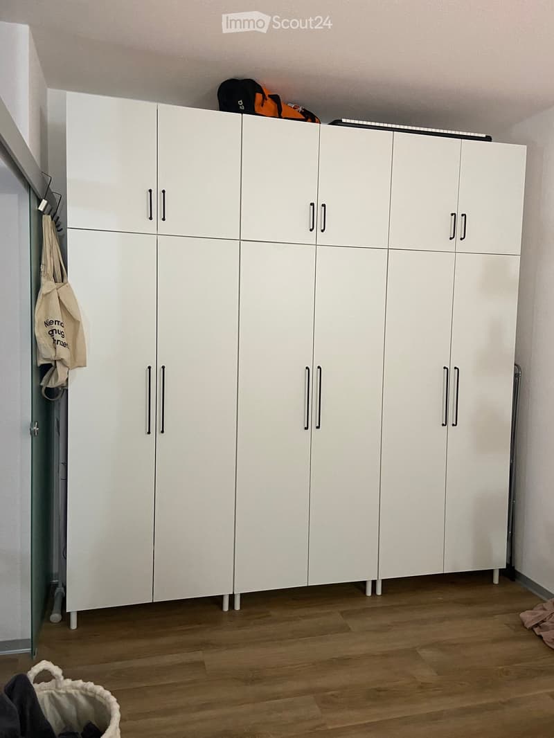 Empty room with white built-in wardrobes, wooden floor, and a few personal items like a bag and a basket on the floor.