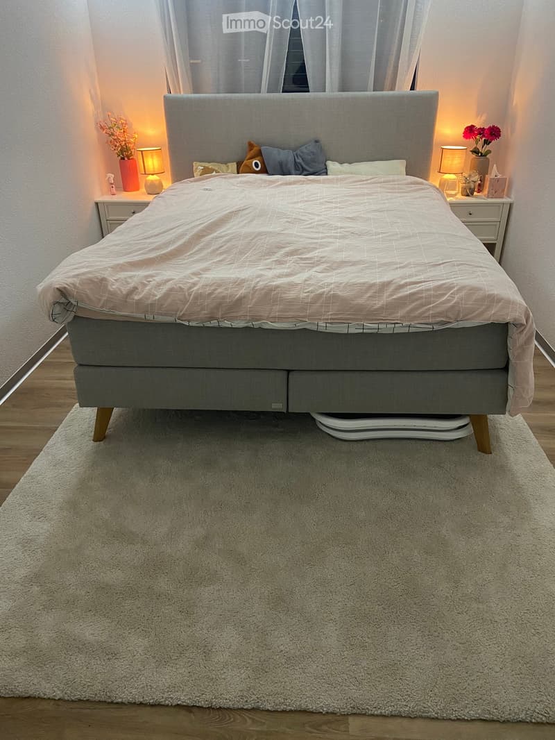 A modern bedroom with a grey upholstered bed, beige duvet, nightstands with lamps, and decorative vases with flowers on either side.