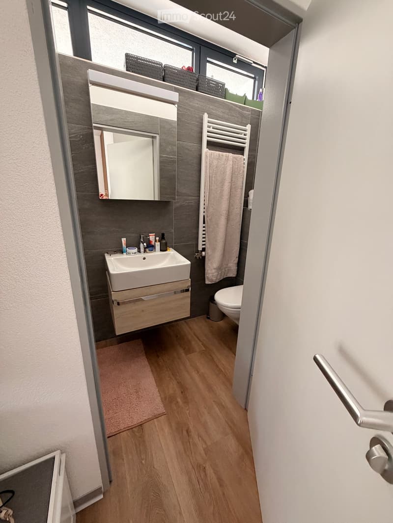 Modern bathroom with wooden flooring, a white sink with a wooden cabinet, a large mirror, a towel on a radiator, and a toilet. The walls are tiled in dark grey, and there is a small window above the sink.