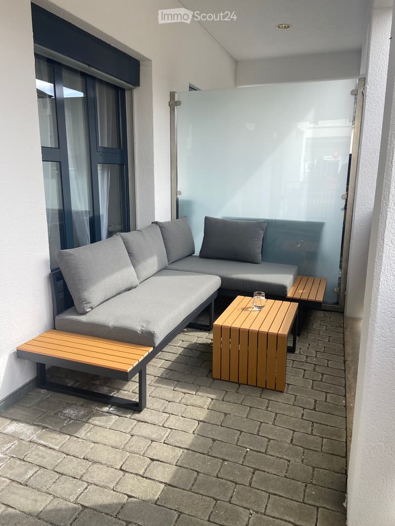 Modern balcony with grey sectional sofa, wooden stools, and a small wooden table holding a glass.