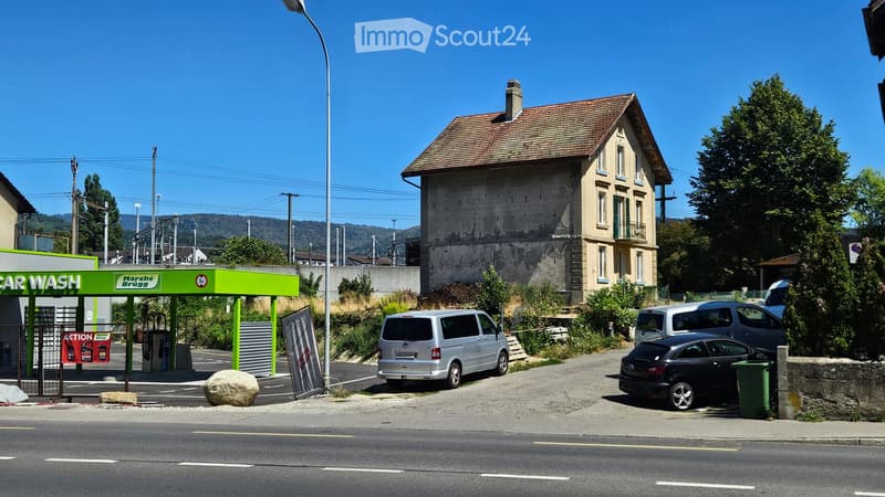 A small building with a tiled roof and a car wash station on the left, with several parked cars and a clear blue sky in the background.
