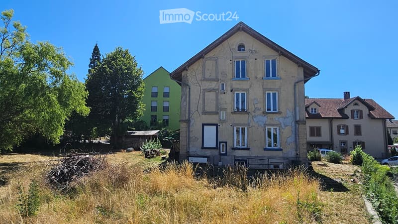 A multi-story residential building with visible wear and peeling paint, surrounded by overgrown grass and trees.
