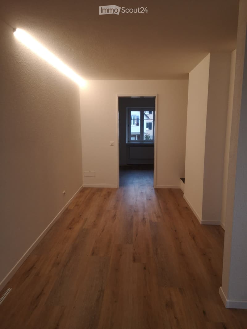 Empty hallway with wooden flooring, white walls, and a doorway leading to another room with a window and radiator visible.
