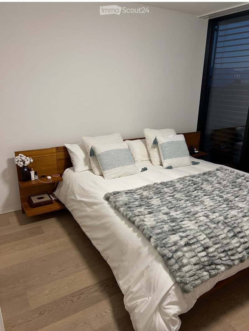A modern bedroom with a large bed, white bedding, decorative pillows, a grey fur throw, a wooden nightstand with a vase and small items, and a large window with blinds.