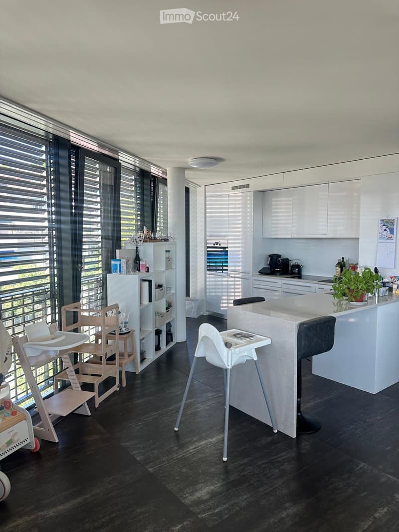 Modern kitchen with white cabinets, dark flooring, large windows with blinds, kitchen island with bar stools, baby chairs, and a shelf with various items.
