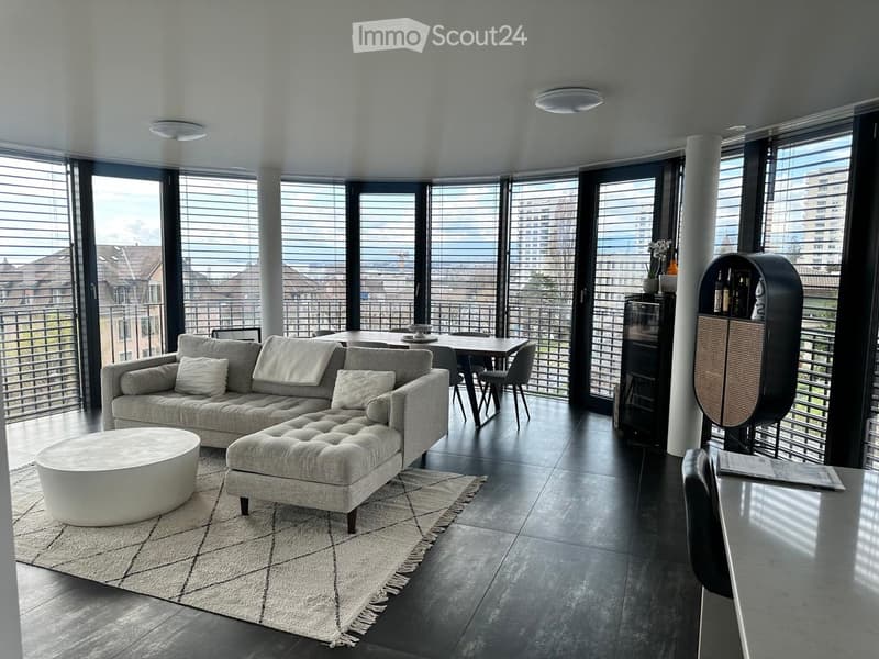 Modern living room with large windows offering a city view, featuring a grey sectional sofa, ottoman, round coffee table, dining table with chairs, and a black cabinet with shelves.