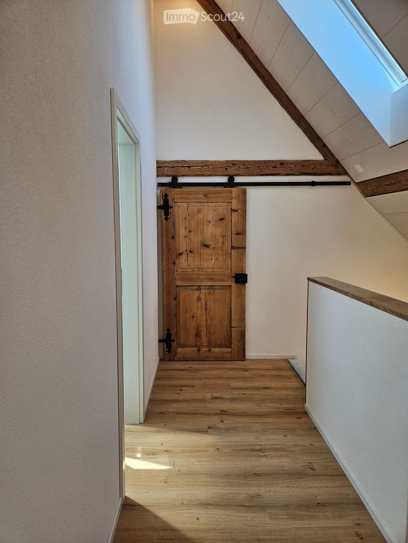 Spacious hallway with wooden flooring, white walls, and a large wooden sliding door leading to another room. A skylight provides natural light, and a wooden railing is visible on the right side.