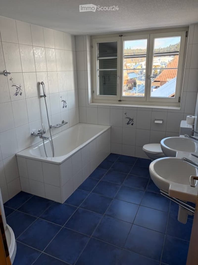 white bathtub, white wall tiles, blue floor tiles, toilet, sinks, window