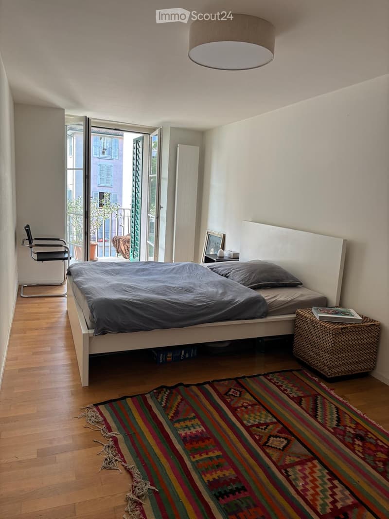 Bedroom with a double bed, nightstand, chair, wicker basket, and a colorful rug on the floor. The room has wooden flooring and large windows with a view of the street.