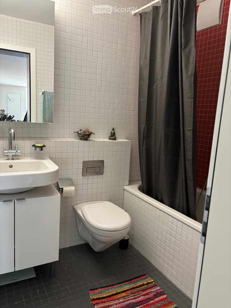 Modern bathroom with white tiles, black floor, sink, toilet, bathtub, mirror, and a colorful rug.