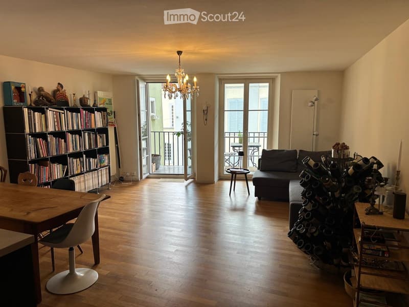 Bright living room with wooden flooring, large windows, and a balcony. Features a sofa, coffee table, bookshelf, wine rack, and chandelier.