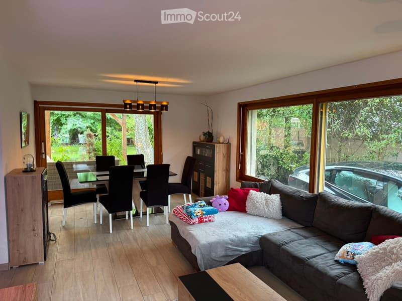 Bright living and dining area with large windows, wooden flooring, a grey sectional sofa, a coffee table, a dining table with chairs, and a cabinet.