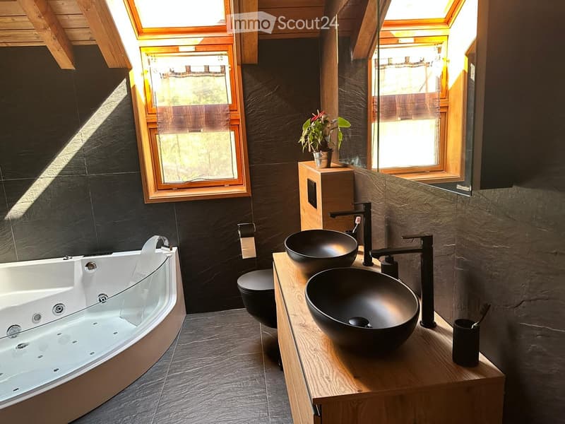 Modern bathroom with a large bathtub, two black sinks on a wooden vanity, a toilet, and a small plant on the vanity.