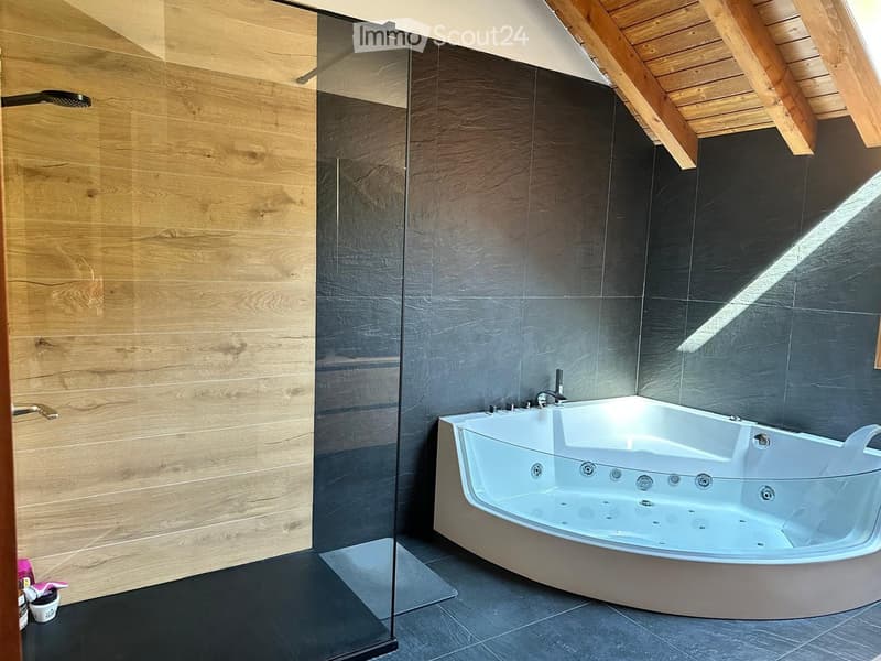Modern bathroom with a large, corner bathtub, sleek dark tiles, and a glass-enclosed shower area. The walls feature a combination of light wood paneling and dark tiles, with a wooden slanted ceiling.