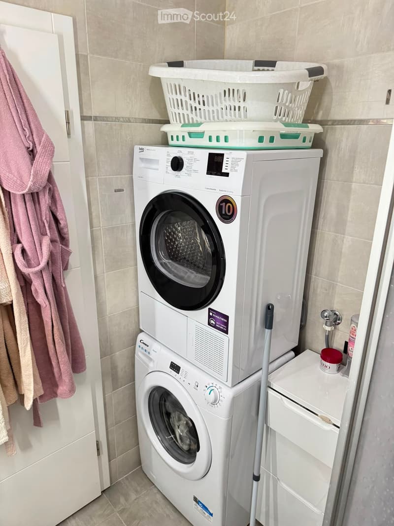 Bathroom with a stacked washing machine and dryer unit, laundry baskets on top, a towel hanging on the wall, and cleaning supplies on a small shelf.