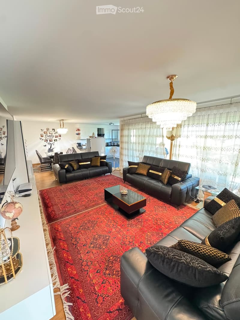 Modern living room with dark leather sofas, a glass coffee table, and a large red patterned rug. The space features a chandelier, wall-mounted TV, and a dining area visible in the background.