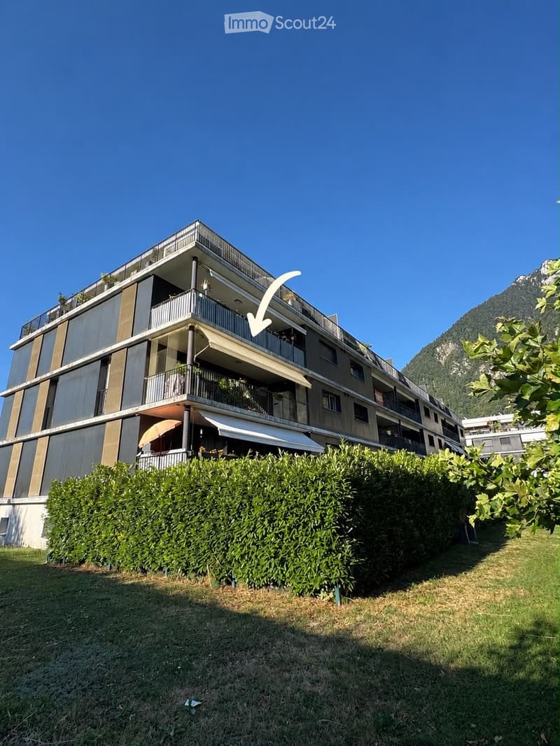 Modern multi-story apartment building with balconies, surrounded by greenery and a mountain view.