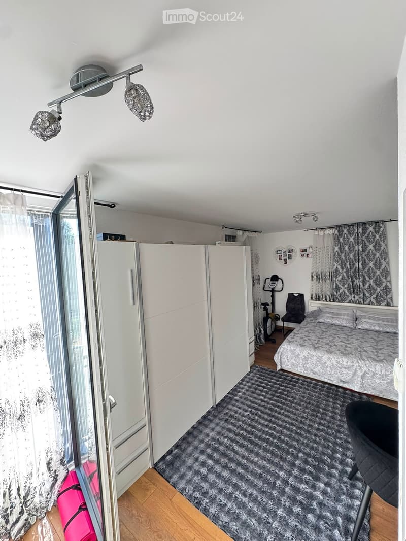 Modern bedroom with a large bed, a grey shaggy rug, a wardrobe, an exercise bike, and a chair. The room features large windows with curtains and decorative wall art.