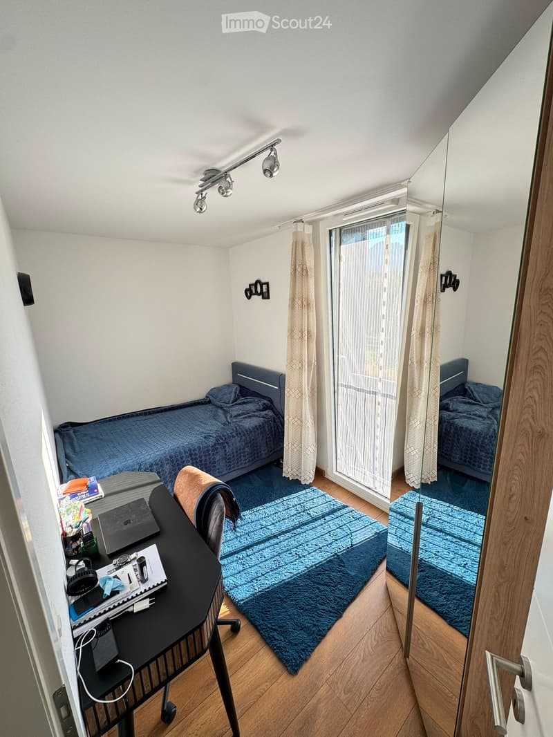 Cozy bedroom with a single bed, desk, chair, and mirror. The room features a blue rug, wooden flooring, and a large window with curtains.