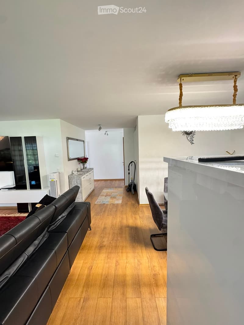 A modern, open-concept living space with a black leather sofa, a wooden floor, a small table with a chair, a mirror, a television, and a decorative flower vase on a side table.