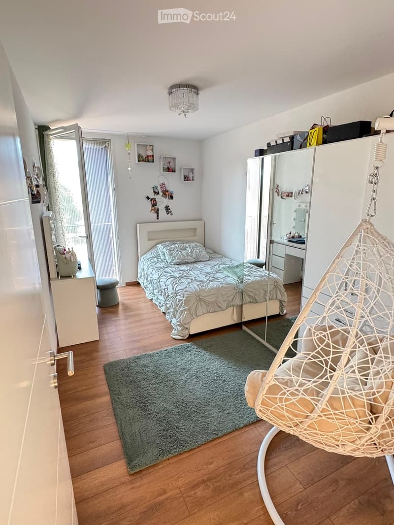 Modern bedroom with a white bed, patterned duvet, wicker hanging chair, large mirror cabinet, and a green shag rug on wooden flooring.