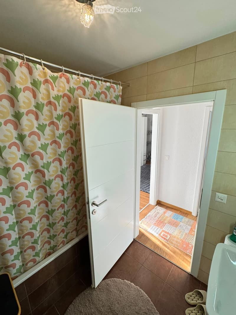 Bathroom with a bathtub, colorful shower curtain, sink, mirror, and a small rug on the floor. A door leads to another room with wooden flooring and a patterned rug.