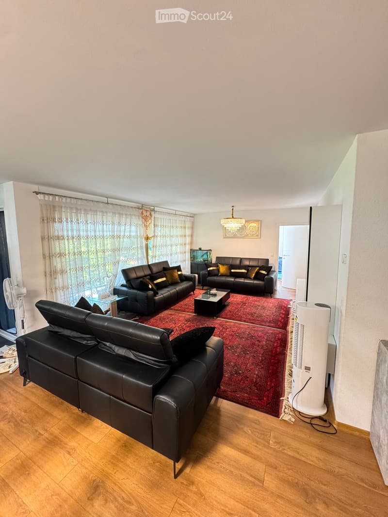 Modern living room with dark leather sofas, a coffee table, a chandelier, and a fan. The room features wooden flooring and large windows with curtains.