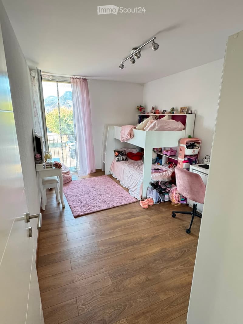 A cozy bedroom with a bunk bed, a small table, a chair, a shelf, and a pink rug on the wooden floor.