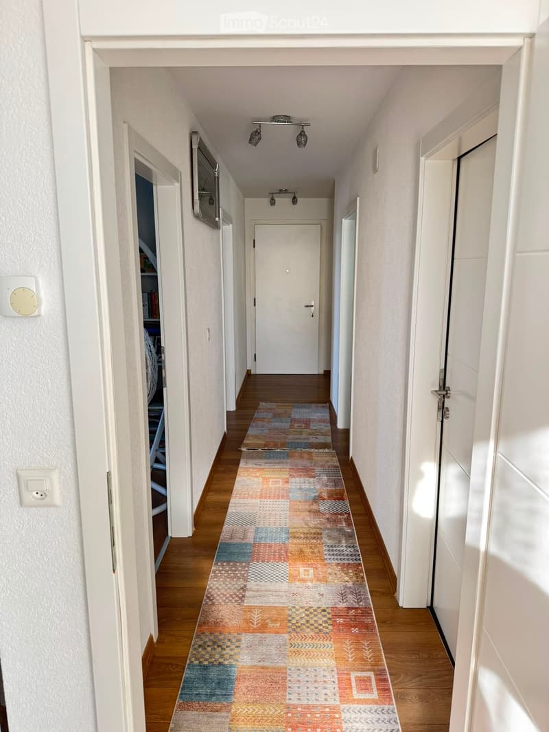 Long hallway with wooden flooring, colorful patterned runner, white walls, several doors, and ceiling lights.