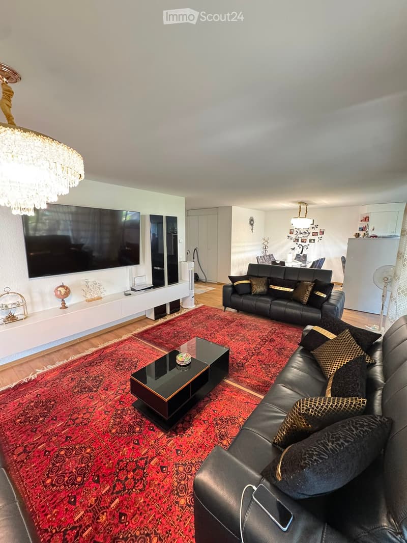 Modern living room with a large red patterned rug, black leather sofas, a glass coffee table, a flat-screen TV, and a chandelier.