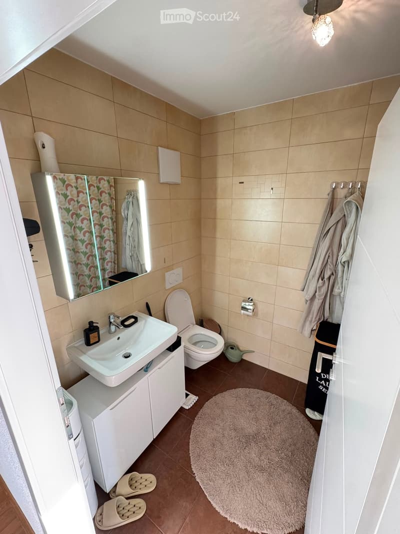 Modern bathroom with beige tiles, white sink, toilet, mirror, and a fluffy rug on the floor. Includes a shower area with a curtain and towels.