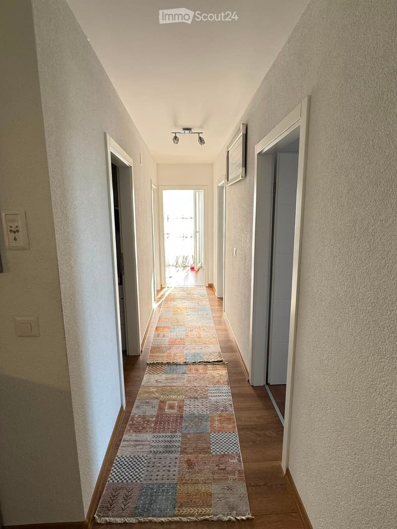 A narrow hallway with wooden flooring, beige walls, and a colorful patterned runner. The hallway features several doors and a ceiling light fixture.