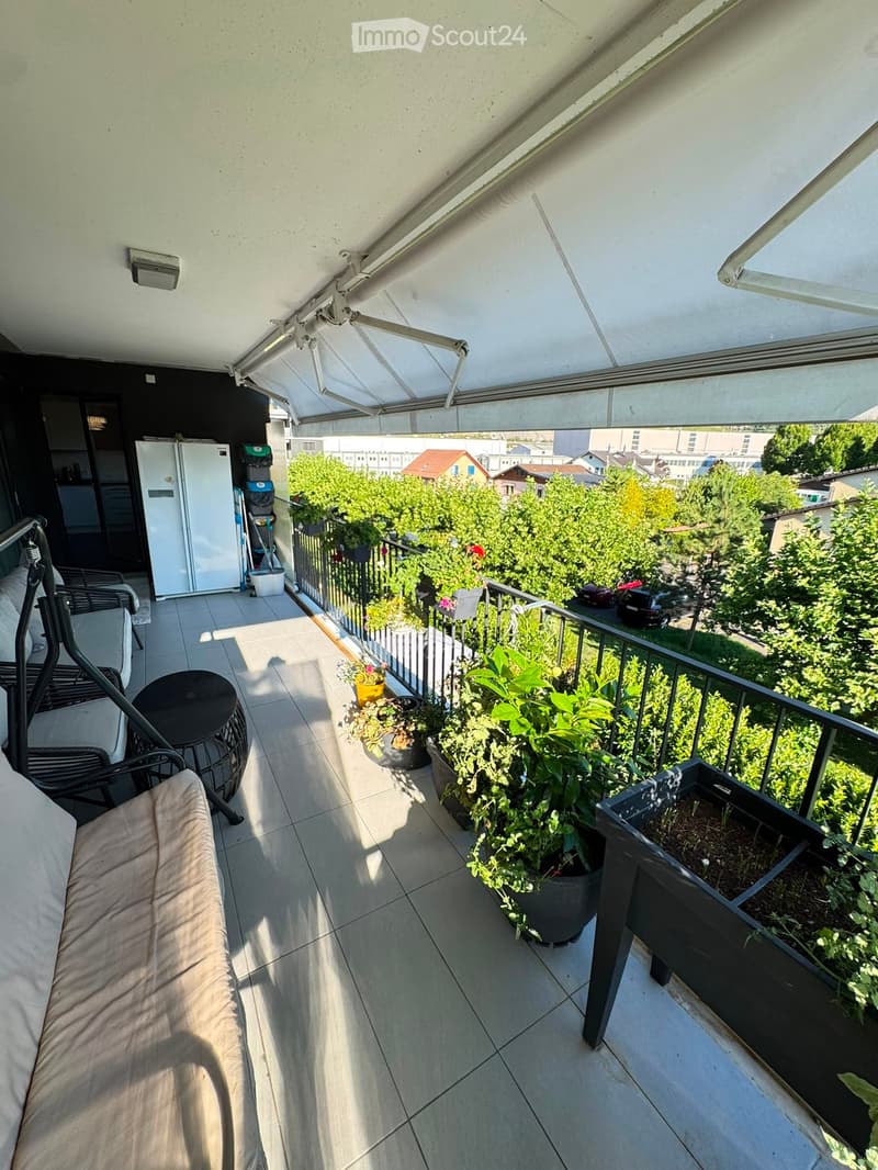 Balcony with tiled floor, sofa, small table, chairs, and several potted plants. The balcony has a retractable awning and overlooks a green area with trees and parked cars.