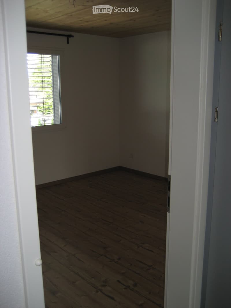 Empty room with wooden flooring and white walls, featuring a small window with blinds and a visible ceiling beam.