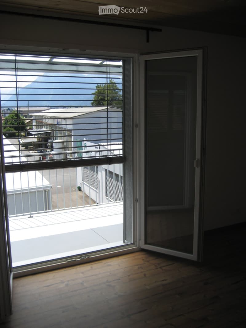 Empty room with wooden flooring and large sliding glass doors leading to a balcony with metal railings and a view of a building and mountains.