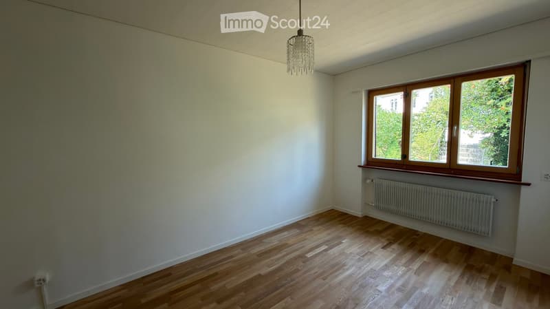 Empty room with wooden flooring, white walls, a large window with wooden frame, radiator, and a hanging chandelier.