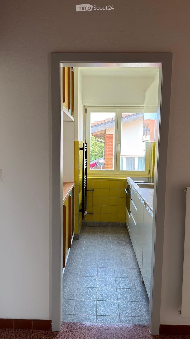 A narrow hallway with tiled floor, white walls, and a door leading to a kitchen area with yellow tiles and white cabinets.