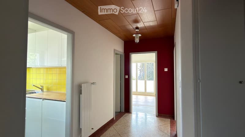 Hallway with wooden ceiling, white and red walls, tiled floor, and access to a kitchen and a room with large windows.
