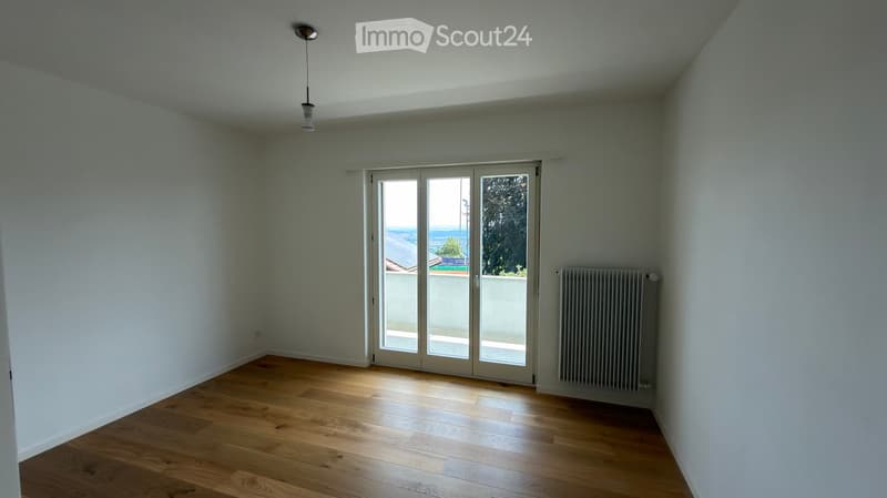 Empty room with wooden flooring, white walls, large sliding glass doors leading to a balcony, and a radiator on one wall.