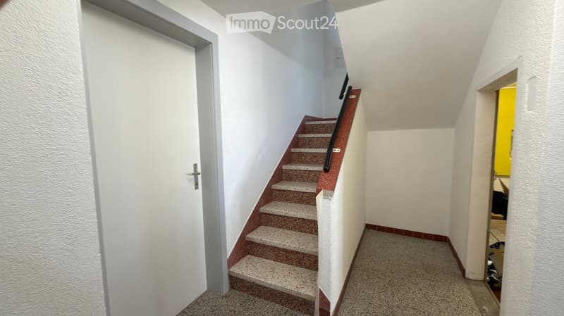 A narrow hallway with a staircase leading upwards, featuring white walls, a grey door on the left, and a small open doorway on the right.