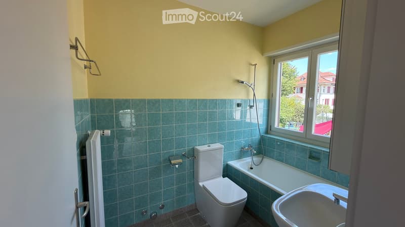 Bathroom with green tiled walls, white toilet, bathtub, sink, radiator, and a window offering a view of the outside.