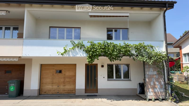 Two-story house with white exterior, wooden garage door, and balcony with green vines. Features multiple windows and a covered entrance area.