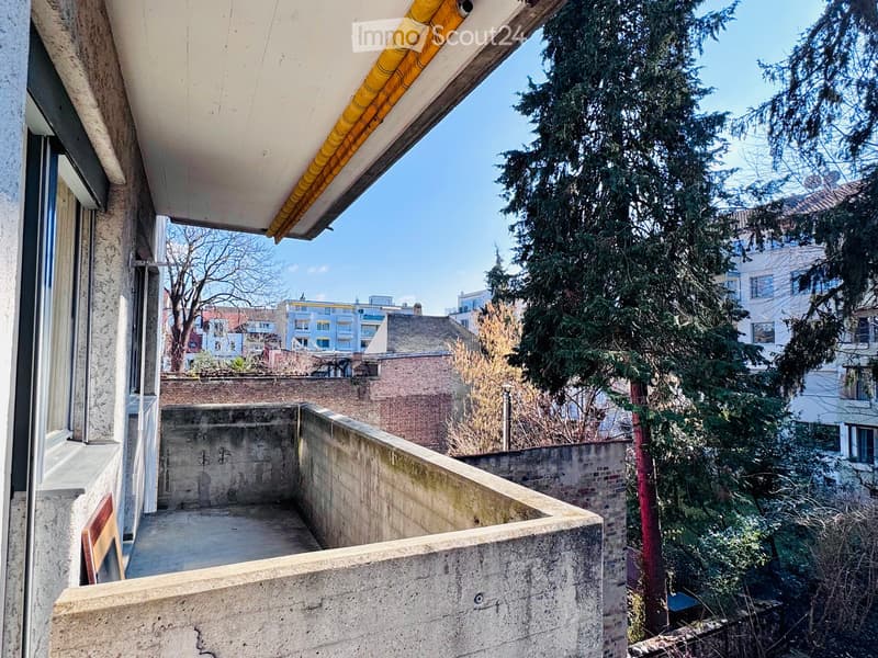 Balcony with concrete floor, low concrete walls, covered by a white roof with yellow trim, overlooking a tree-lined urban area.