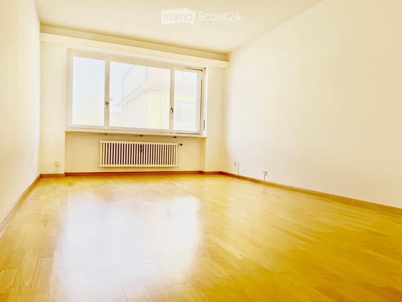 Empty room with wooden flooring, white walls, large window with radiator below, and visible electrical outlets.