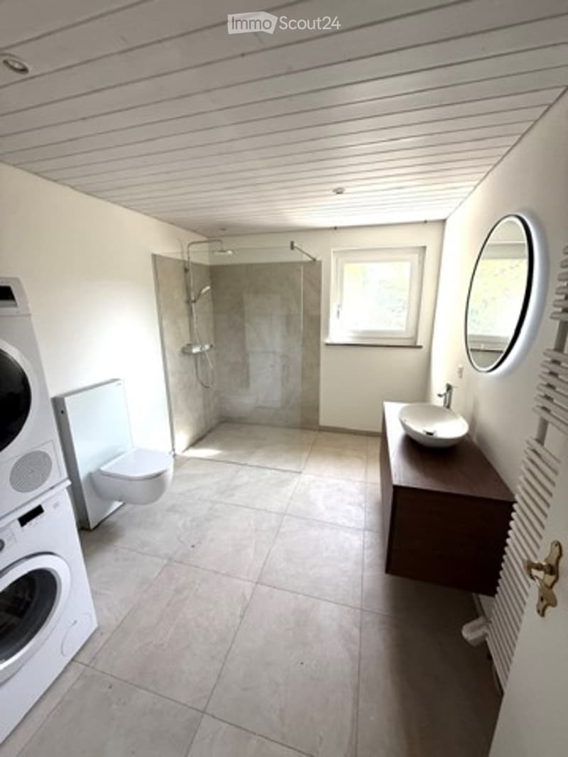 Modern bathroom with a shower, toilet, sink, and washing machine. Features a large mirror, tiled floor, and a radiator.