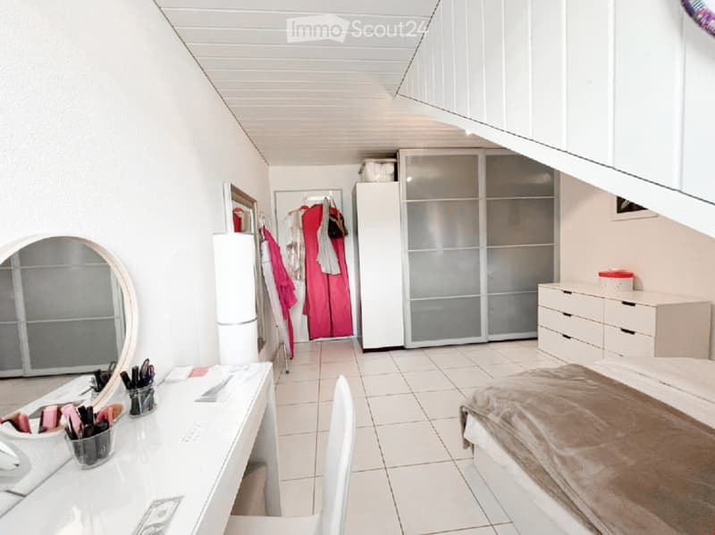 Modern bedroom with white walls, tiled floor, bed with brown blanket, large wardrobe with mirrored doors, dresser, desk with makeup items, and a mirror on the wall.