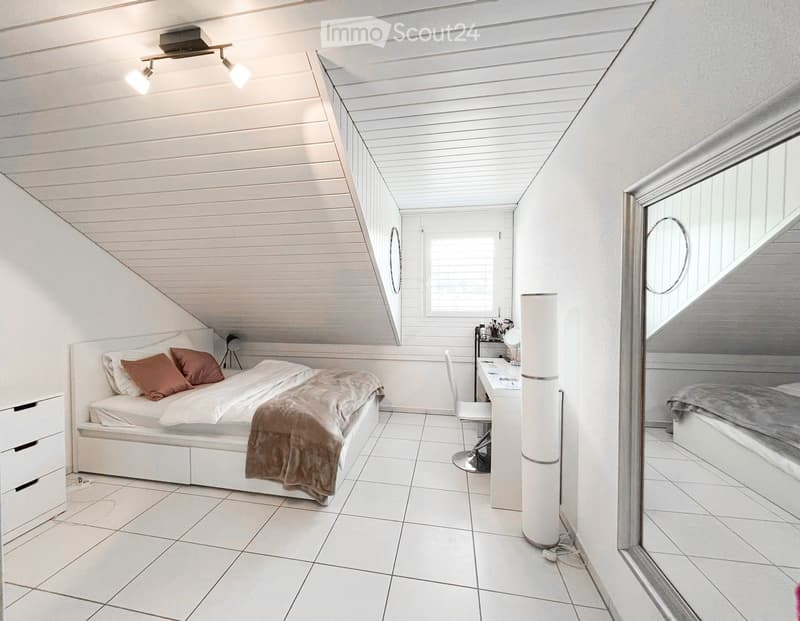 Modern bedroom with white walls, slanted wooden ceiling, tiled floor, bed with pillows and blanket, desk with chair, and a large mirror on the wall.