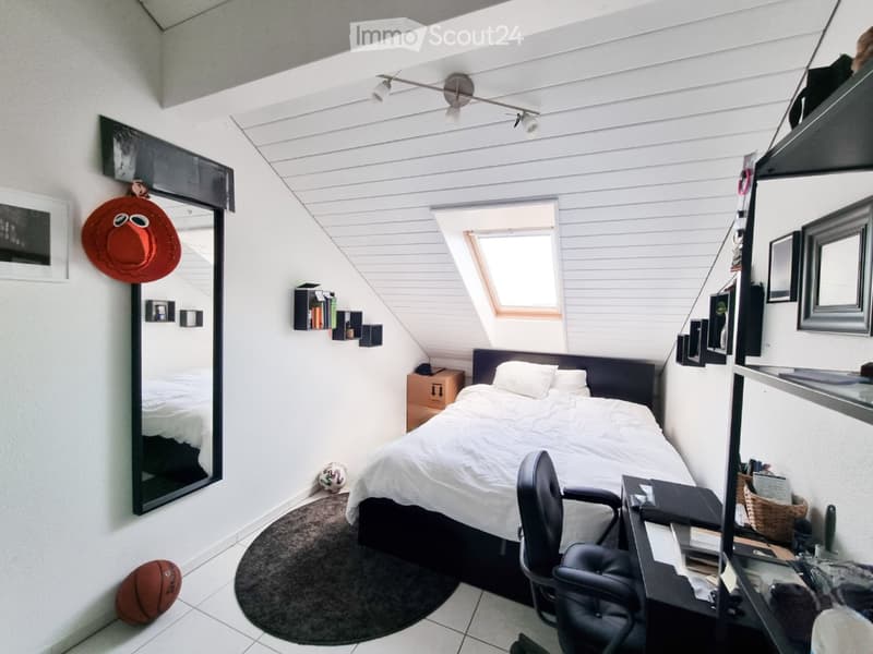 Cozy attic bedroom with slanted ceilings, a bed with white bedding, a desk with a chair, shelves, a mirror, and decorative items including a hat and a ball.