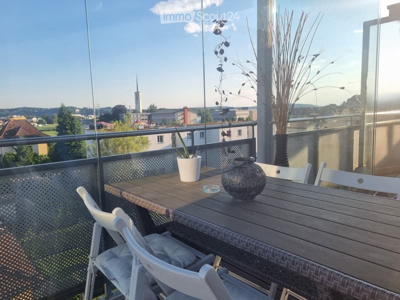 Balcony with a wooden table, white chairs, decorative vases, and potted plants, overlooking a cityscape with trees and buildings.
