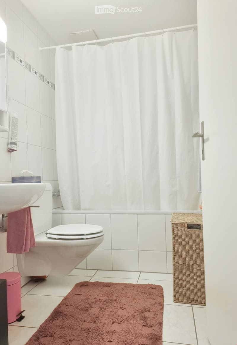 Modern bathroom with white tiles, bathtub, toilet, sink, shower curtain, and a wicker basket on the floor.